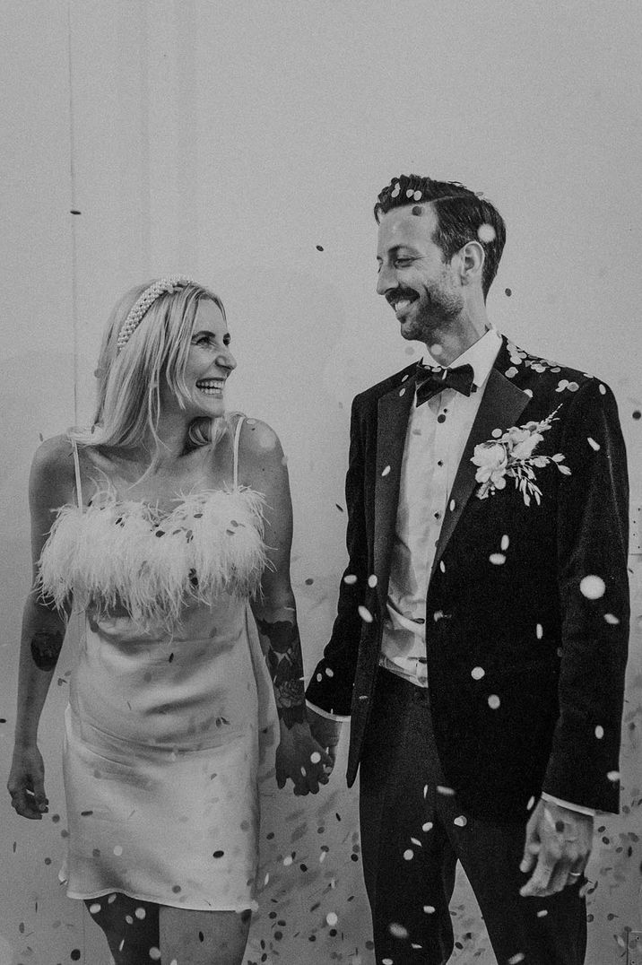 Bride wearing a feather white and short dress with a pearl headband smiling with the groom in black tie for London wedding 