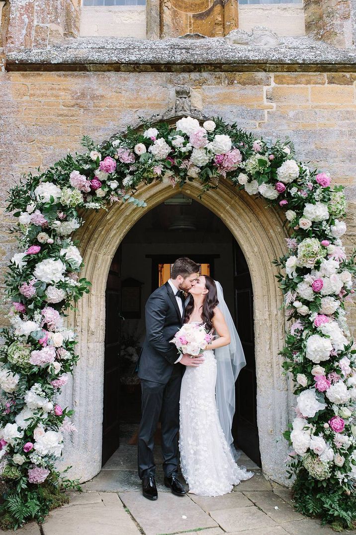 Couple stand under doorway surrounded by pink and white wedding flowers