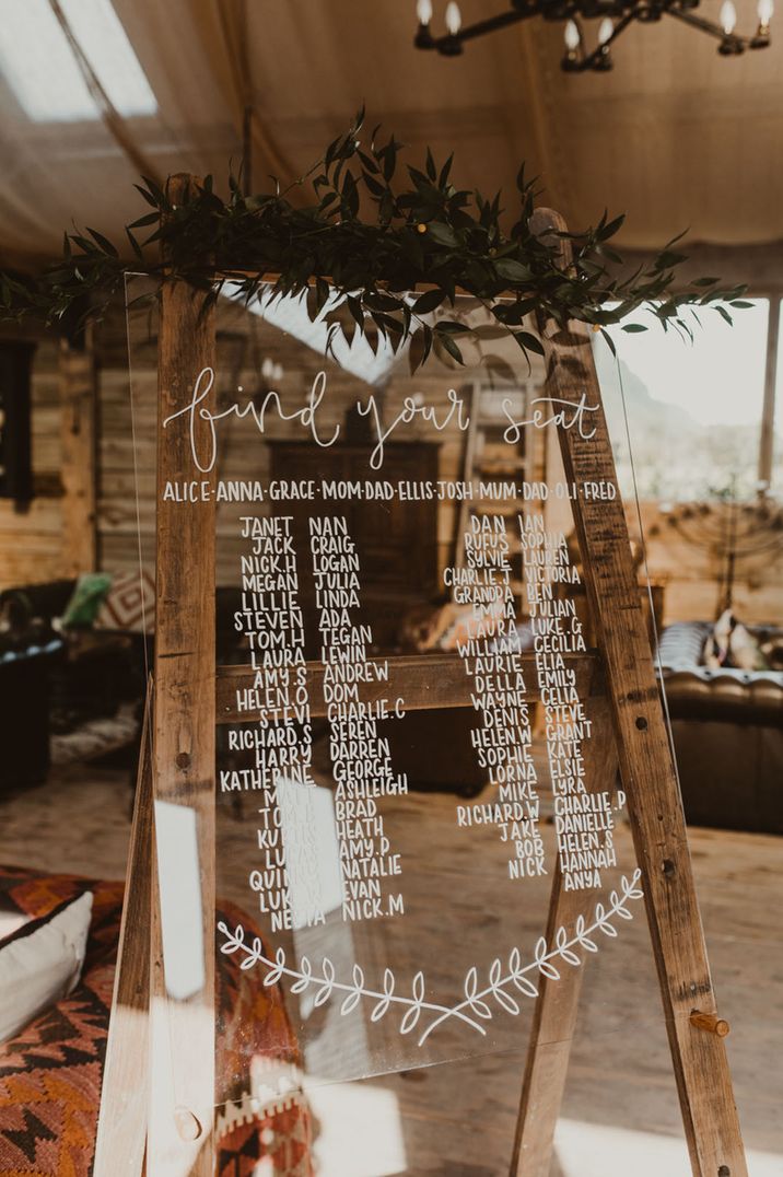 Clear acrylic table plan with white font and green foliage decor on rustic wooden easel 