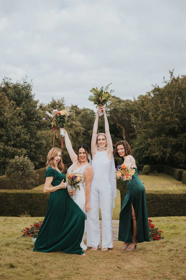 Bride in sleeveless bridal jumpsuit with cape standing with bridesmaids in bottle green velvet bridesmaid dresses 