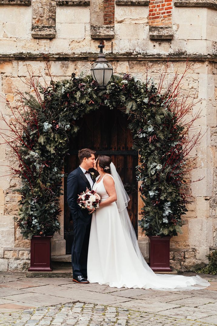 The bride and groom kiss at the entrance to their rustic barn wedding venue decorated with a stunning festive seasonal flower arch 