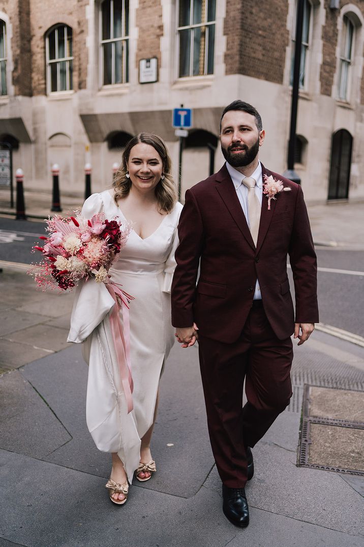 Bride carrying pink and red summer wedding bouquet with red dyed bunny grass and dried palm laves with the groom wearing a matching pink dried flower buttonhole 