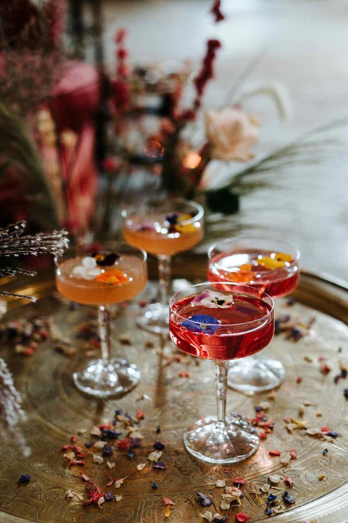 Four colourful wedding cocktails with edible flower toppers in cocktail glasses on wooden slate