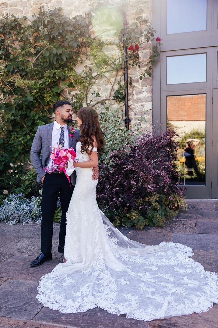 The bride stands in an off the shoulder lace wedding dress with long train posing with the groom in a grey suit 