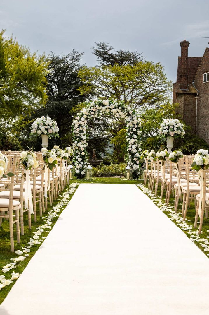 Outdoor area of Farnham Castle wedding venues with large white garden rose and foliage floral arch and white garden rose aisle flower decorations and petals 