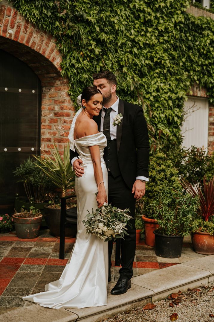 The bride in a classic white dress stands with groom in black tuxedo for their couple portrait together 