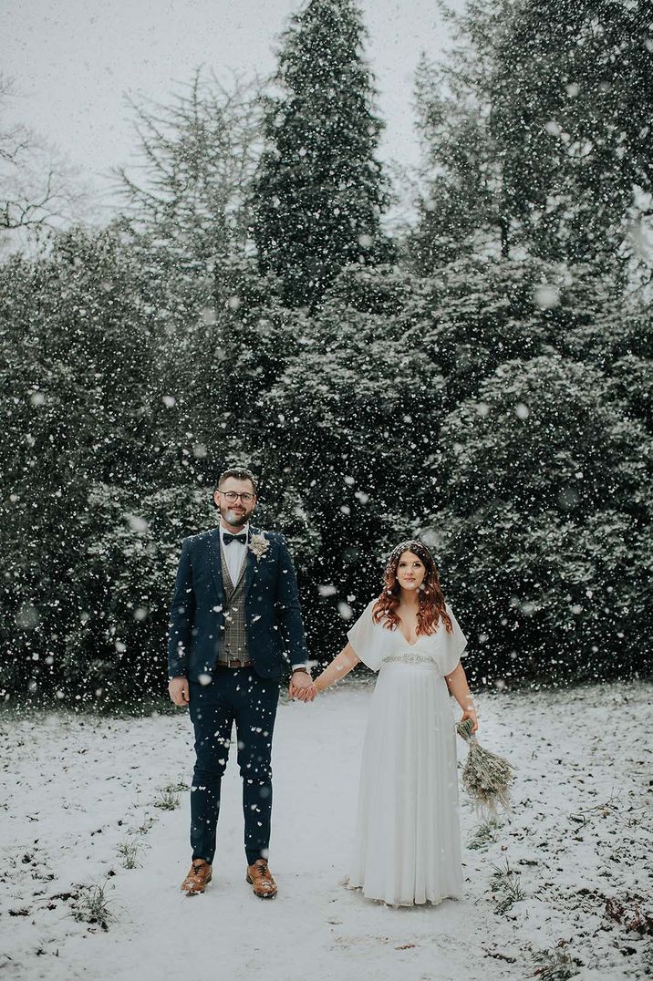 Bride and groom stand holding hands for their winter wedding in the snow 