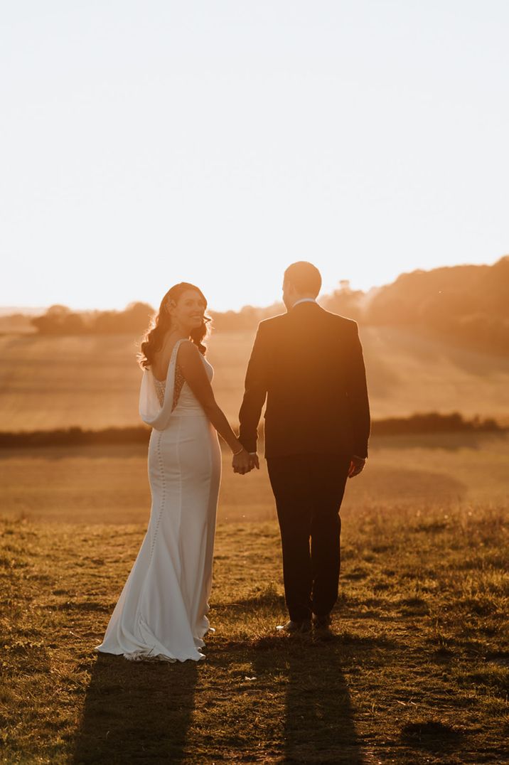 Bride in sparkly draped back wedding dress walking hand in hand with the groom during sunset 