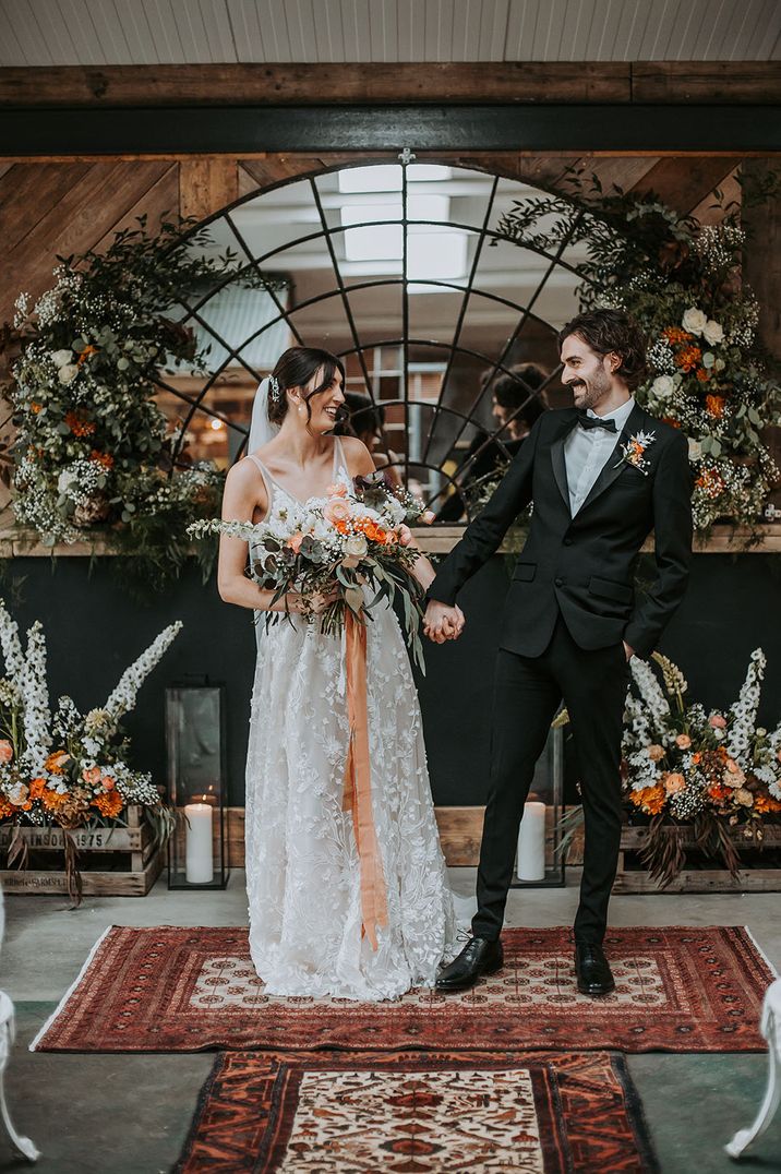 Bride in floral lace wedding dress standing with groom in a black tuxedo at Ghyll Barn Lake District wedding venue 