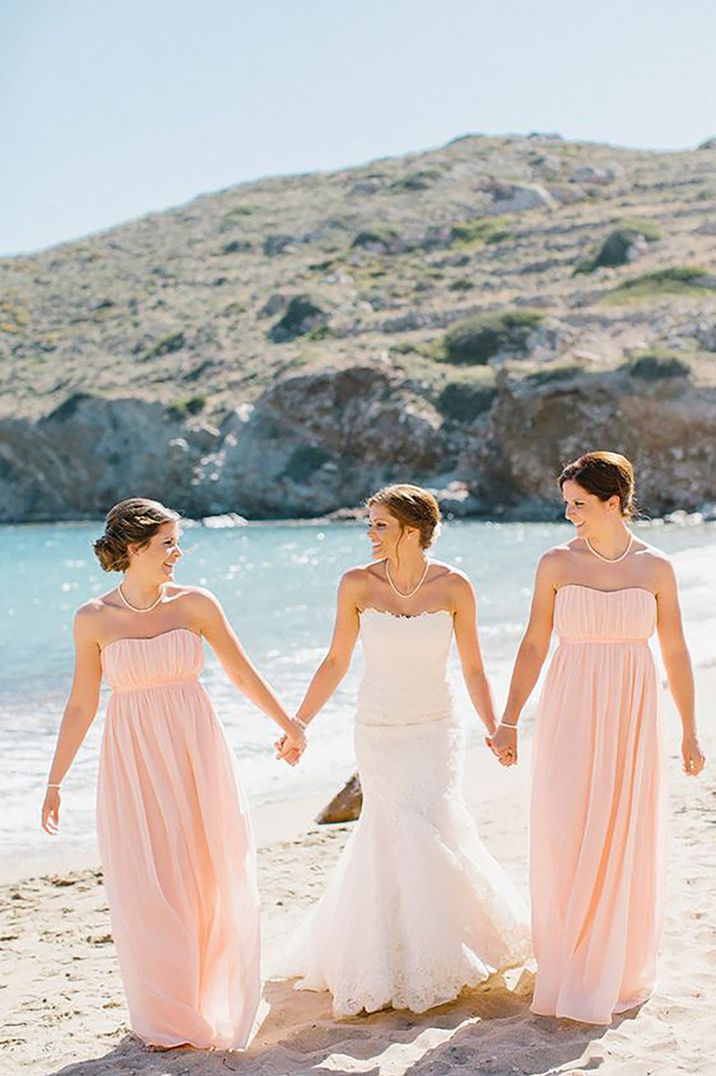 Bride holding hands with her two bridesmaids on a beach wearing peach bridesmaid dresses for greek destination wedding by Anna Roussos Photography