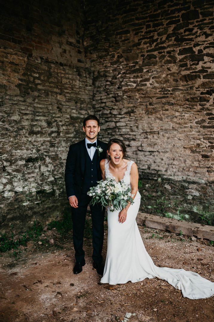 Groom in a navy suit and bow tie laughing with his bride at Dewsall Court with the bride in a fitted wedding dress with lace bodice holding a white and green wedding bouquet 