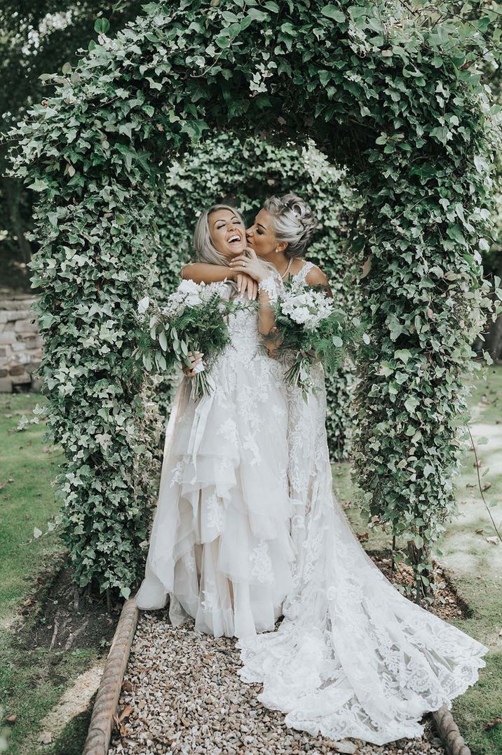 Couple at LGBTQI+ wedding holding white and green bouquets embracing under trees 