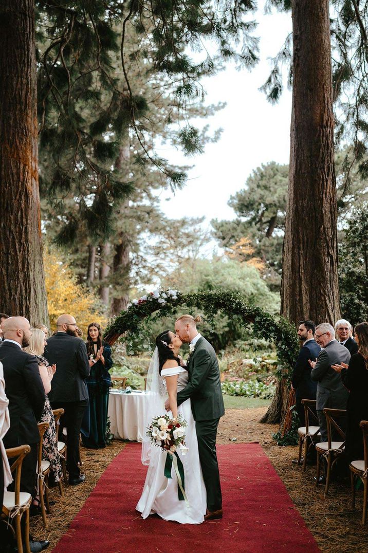 Bride in off the shoulder wedding dress kissing groom in classic grooms suit at Royal Botanic Garden Edinburgh in front of wedding foliage arch 