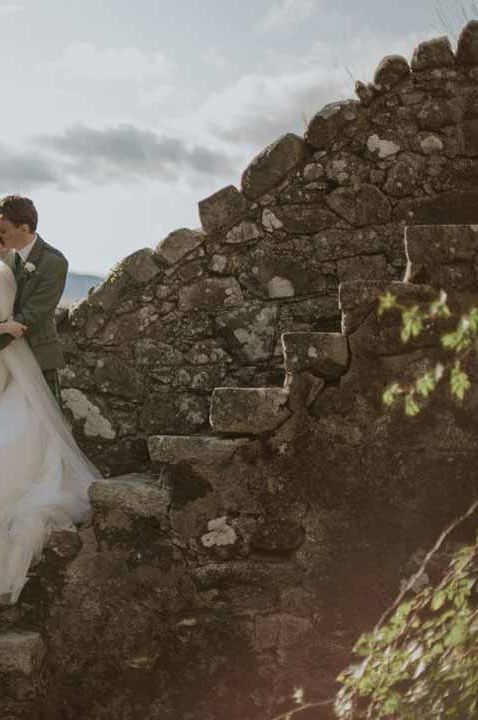 Bride in dramatic sleeveless princess wedding dress kissing groom in classic grey grooms suit on staircase at Barra Castle wedding venue Scotland