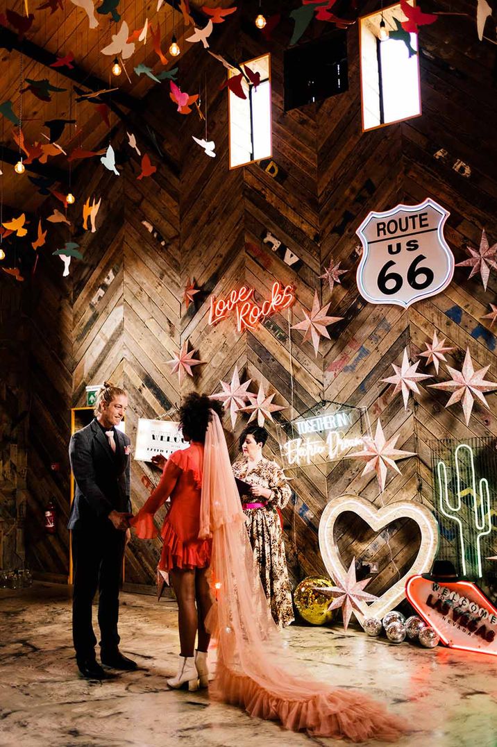 Bride in alternative short red wedding dress with pink bridal veil standing with groom in classic black tux at The Canary Shed wedding venue with lots of neon wedding signs and suspended paper birds 