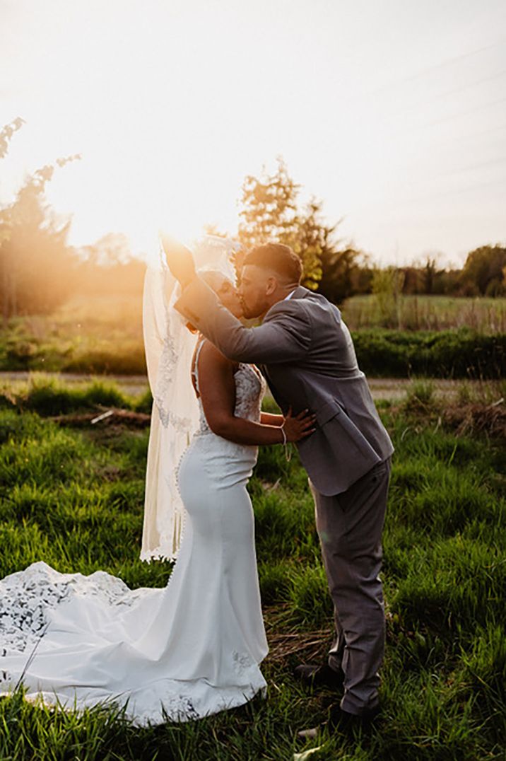 The groom in a light grey suit removes the bride's veil as they kiss during the sunset 