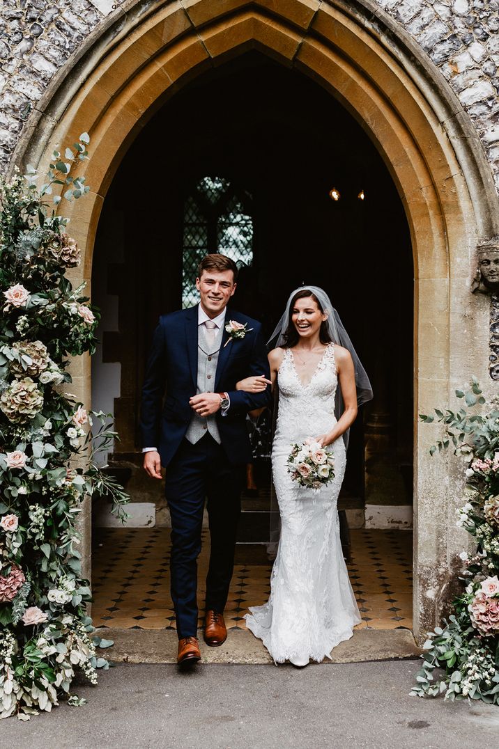 Groom in a navy suit with grey waistcoat and pink tie walking out of the church with his bride in a lace wedding dress