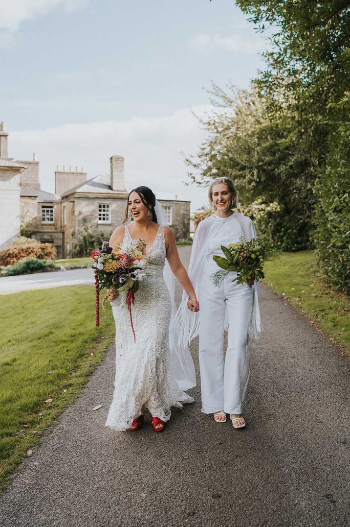 Lesbian wedding with two brides in a lace wedding dress and bridal jumpsuit and cape holding a brightly coloured wedding bouquet 