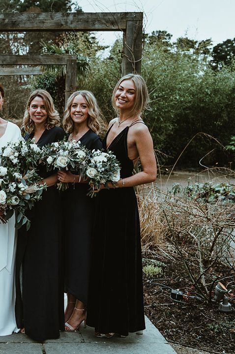 Bride stands with her bridesmaids who wear black bridesmaid dresses in different styles whilst holding white floral bouquets at the Le Petit Chateau