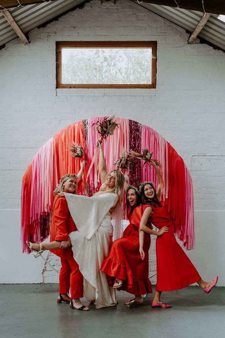 Bridal party in bold red mismatched bridesmaid outfits, white batwing wedding dress and pink steamer decorations - how to save for your wedding