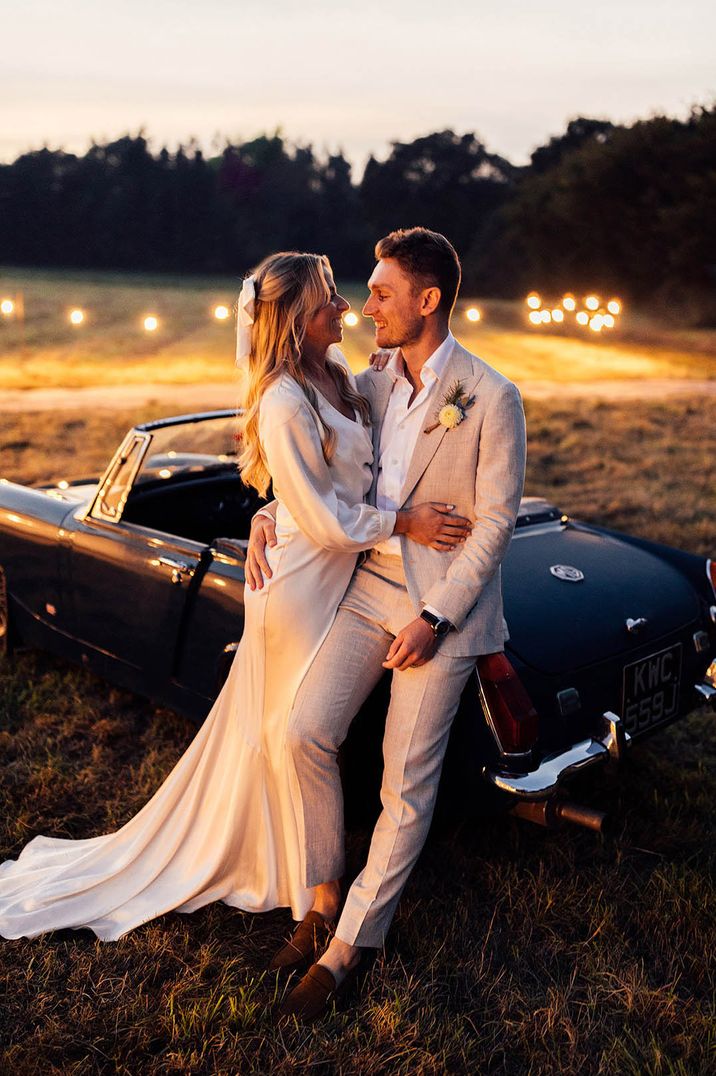 Bride wearing a long sleeve satin wedding dress leaning against the dark blue wedding car with the groom 