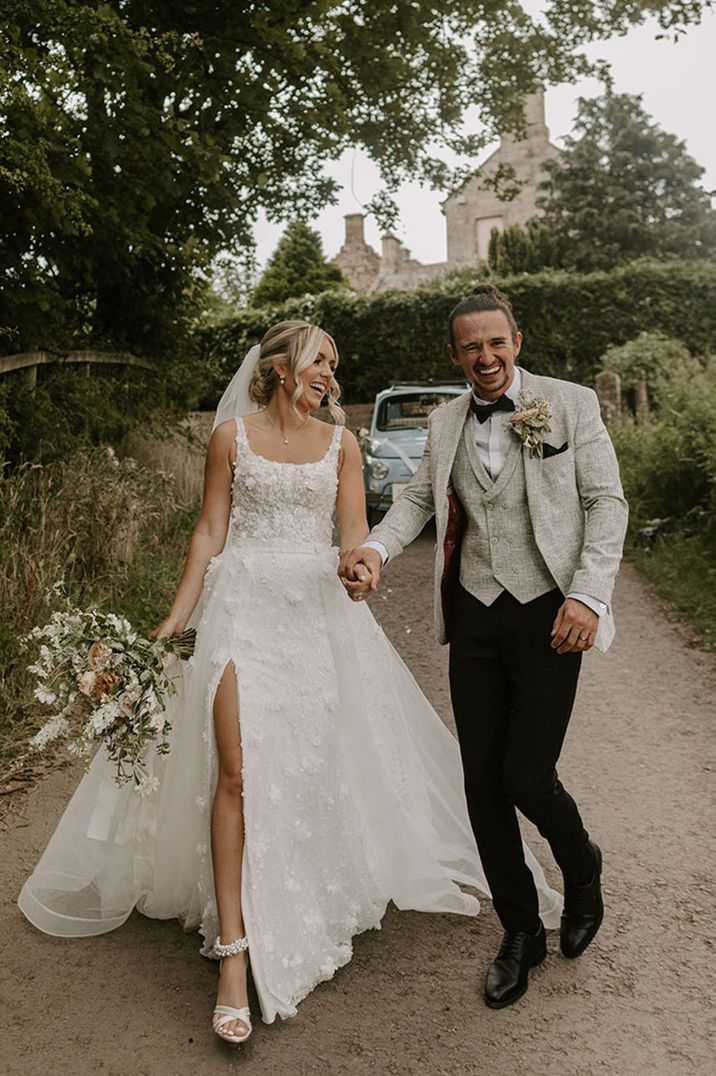 Bride in Eleganza Sposa wedding dress with groom in grey tweed waistcoat and suit jacket with black bow tie in front of vintage blue car 