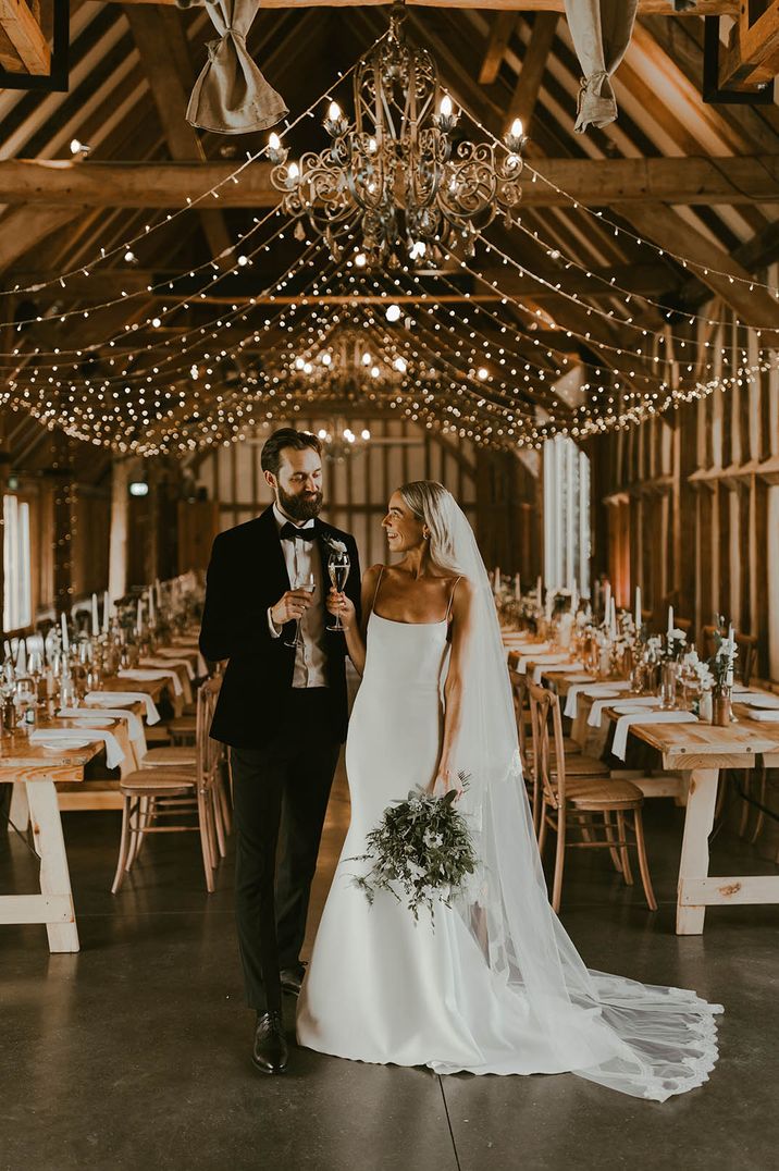 Bride in subtle mermaid wedding dress with thin straps with the groom in a black tuxedo at a barn wedding venue with fairy lights 