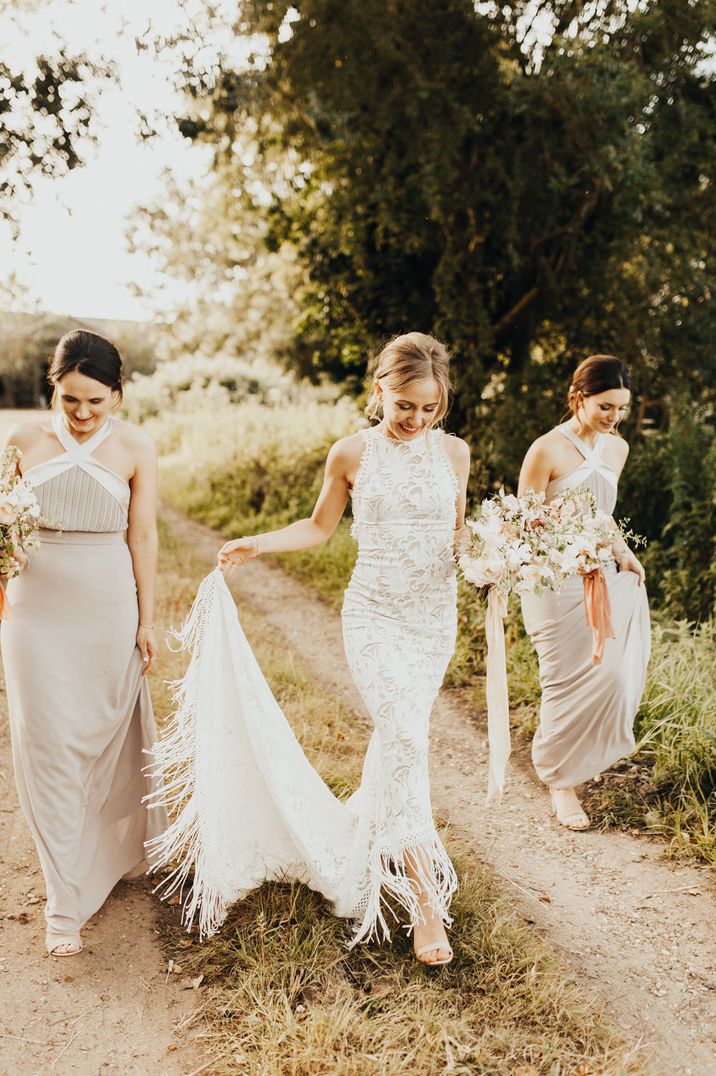 Bride in a fitted Grace Loves Lace Edie fringe wedding dress walking with her bridesmaids by her side in beige halter neck dresses 