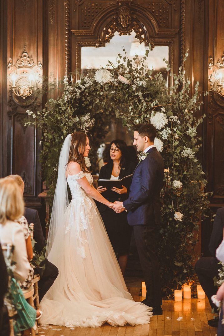 Bride in a strapless Martina Liana wedding dress exchanging vows with her husband in a navy suit in front of a green foliage and white flower arch 