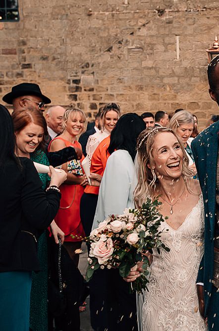 Family portrait confetti moment with groom in a check waistcoat and bow tie holding his flower girl daughter in his arms and embracing his wife in a lace wedding dress