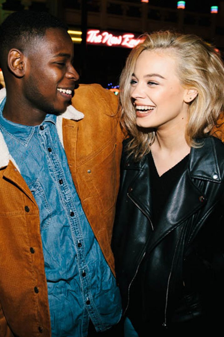 Woman in leather jacket and man in denim shirt smile together at night time 