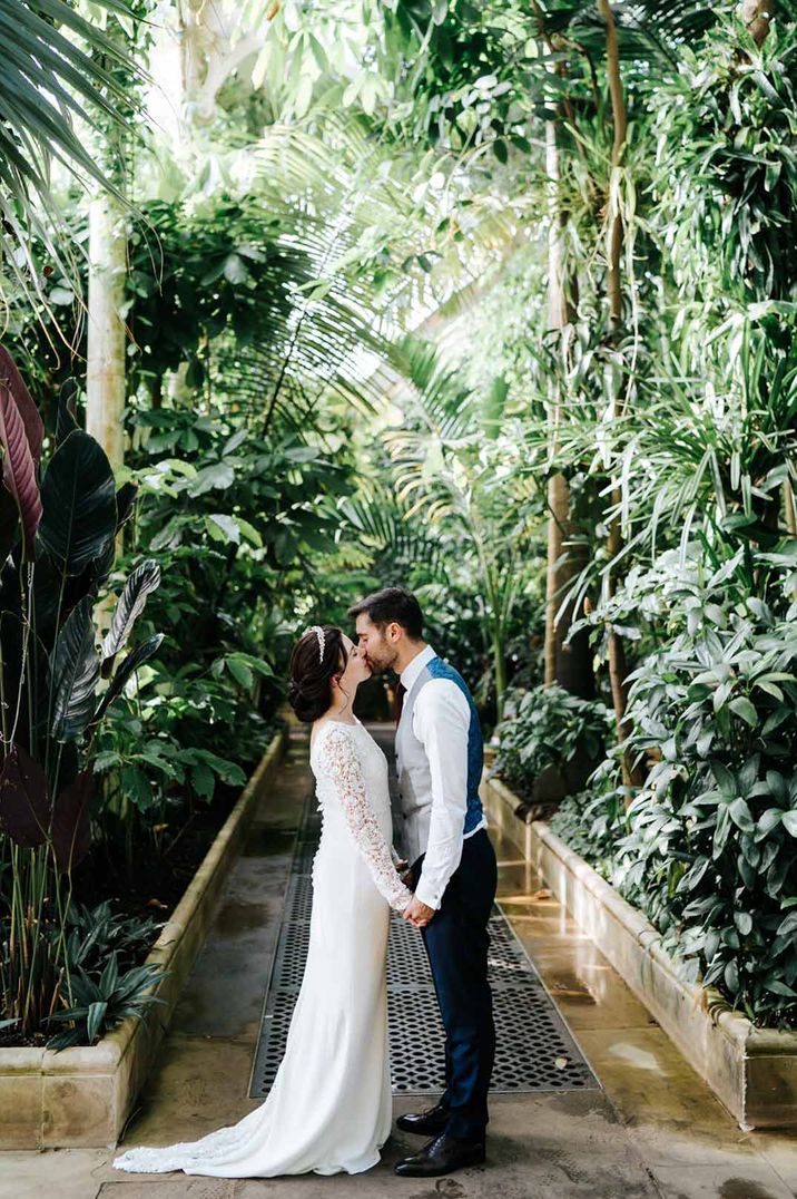 Bride in lace long sleeve wedding dress and groom in grey waistcoat kissing in Kew Gardens London glasshouse wedding venue 