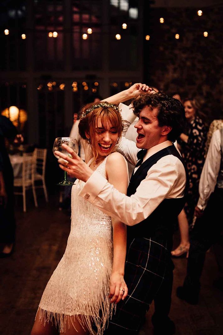 Bride in short fringed wedding dress and ethereal flower crown dancing with groom in waistcoat and white shirt at Oxnead Hall 