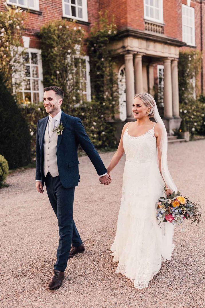 Bride in strappy lace wedding dress holding mixed neutral bouquet with eucalyptus holding hands with groom in three piece blue and grey suit at Iscoyd Park wedding venue 