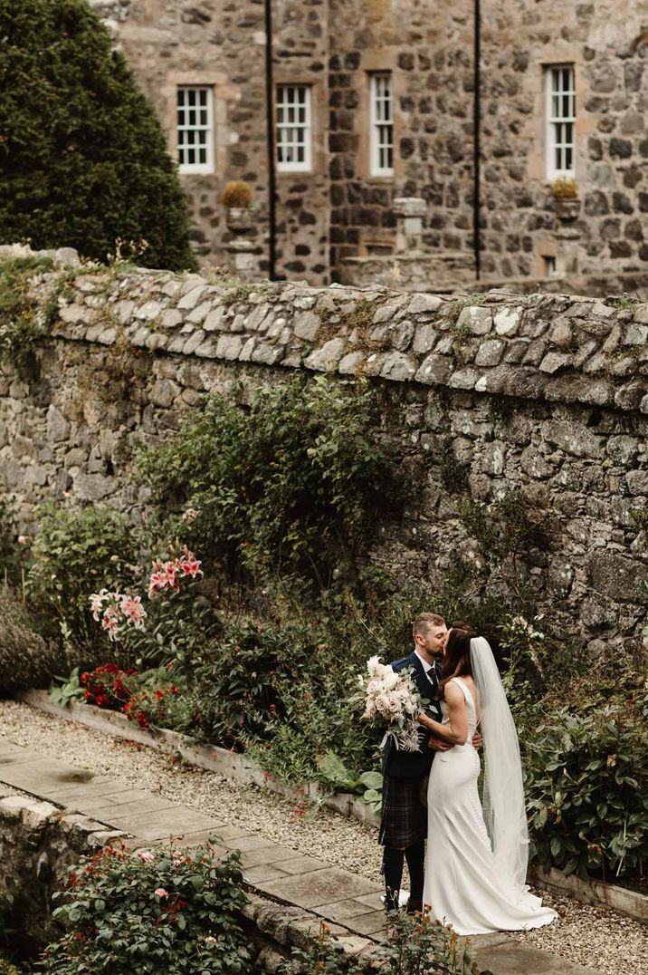 Bride in satin slip wedding dress with church length veil kissing groom in classic black grooms tuxedo at Barra Castle wedding venue Scotland 