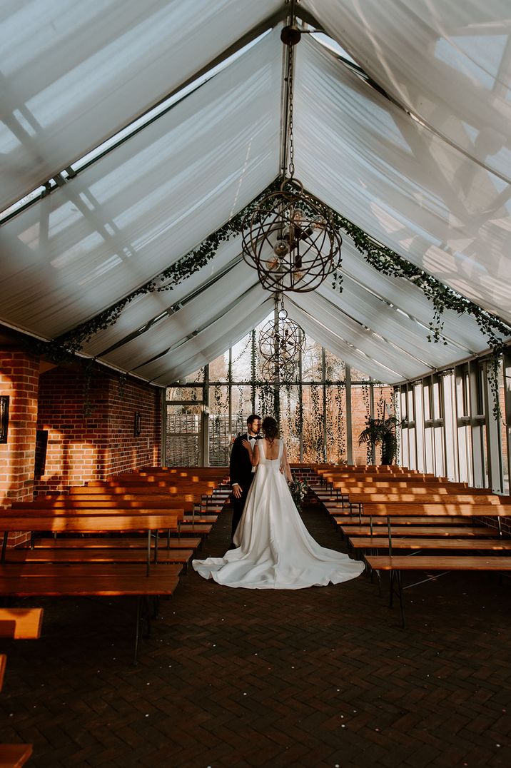 The bride and groom stand in the glasshouse decorated with white drapery and foliage 