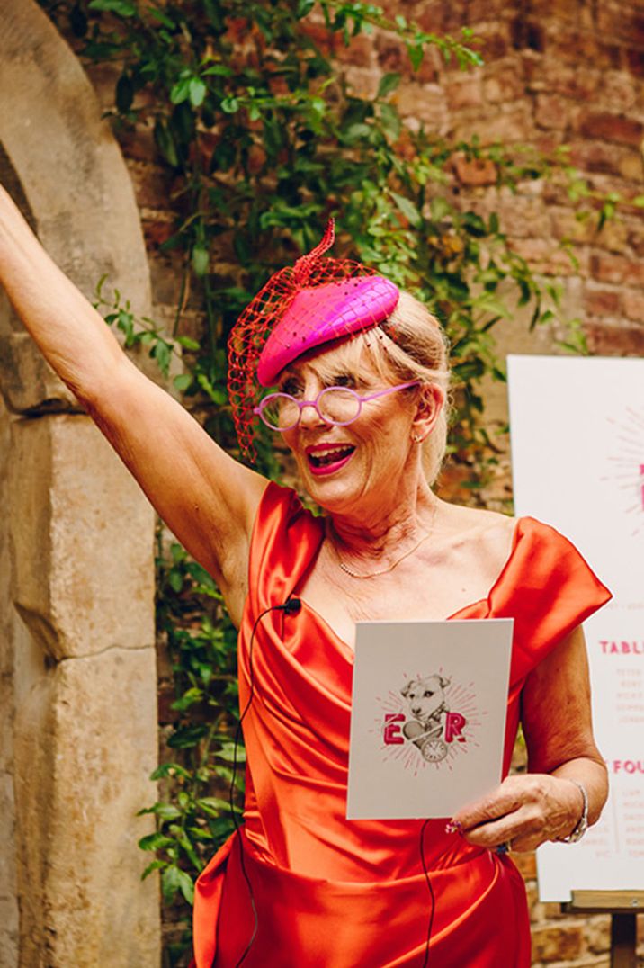 Mother of the bride in orange off the shoulder dress and hat with her hands in the air performing mother of the bride speech 