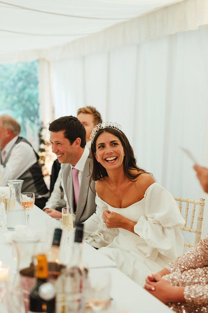 Bride in off shoulder dress and tiara crown listening to maid of honour speech