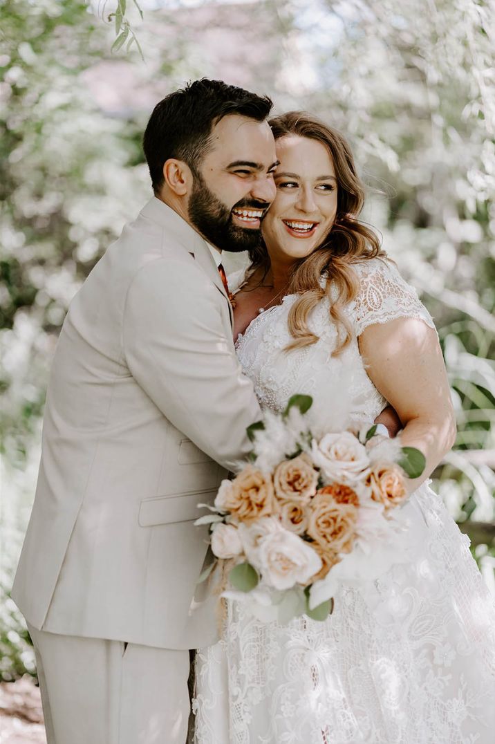 Groom in beige wedding suit hugging the bride tightly wearing a lace wedding dress holding a white and orange rose bouquet 