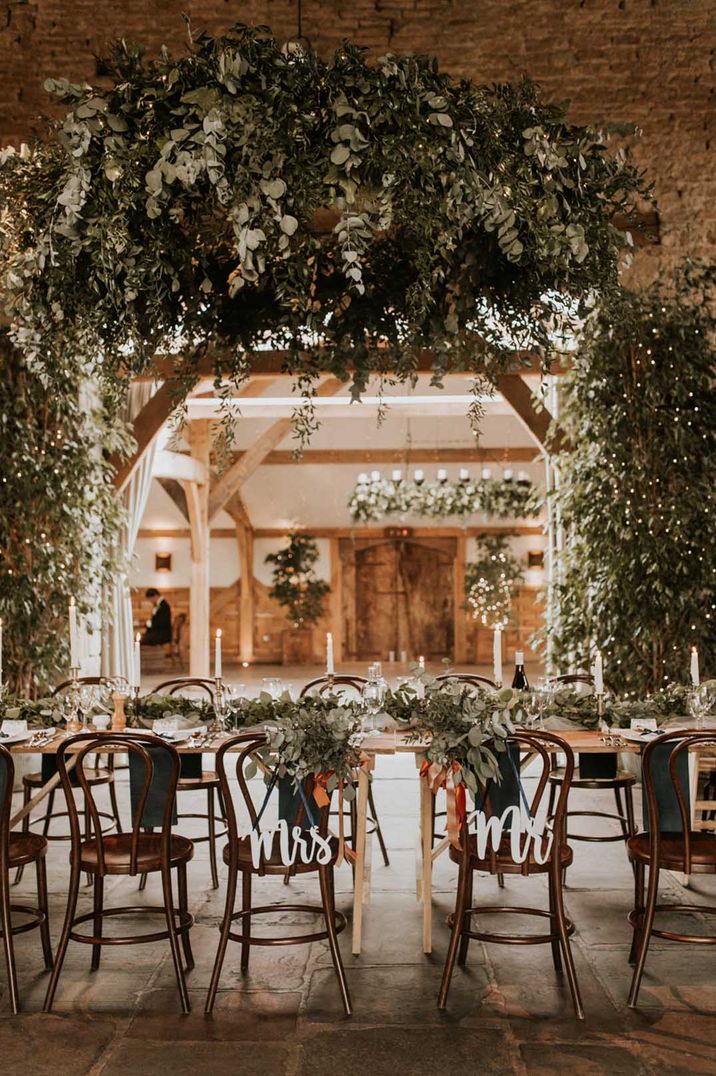 Neutral rustic wedding tablescape in Cripps Barn wedding venue reception room with suspended eucalyptus and foliage decor, Mrs and Mr wooden signs hung on the chairs, tapered candles and fairy lights 