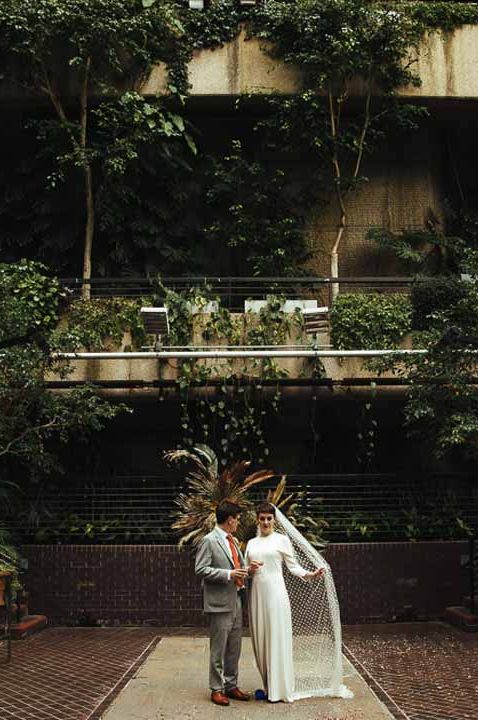 Bride in long sleeve wedding dress with church length veil and groom in grey three piece suit standing in Barbican Conservatory, showing how to book a wedding venue 