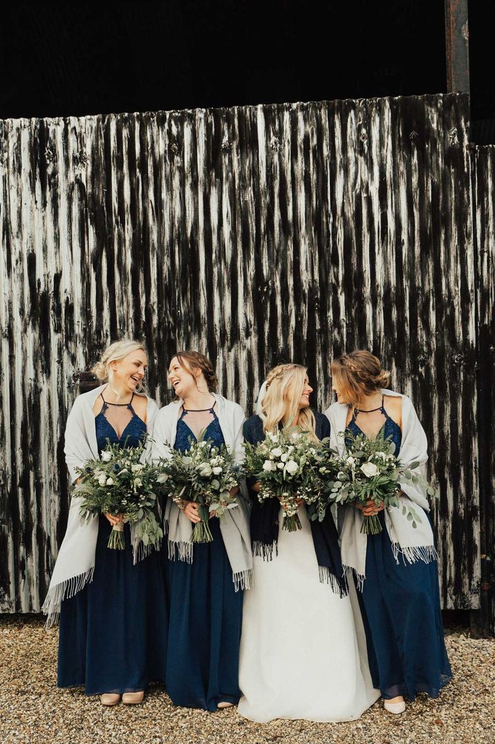 Bride in wedding dress with black shawl standing with bridesmaids in strappy navy bridesmaid dresses and white shawls 