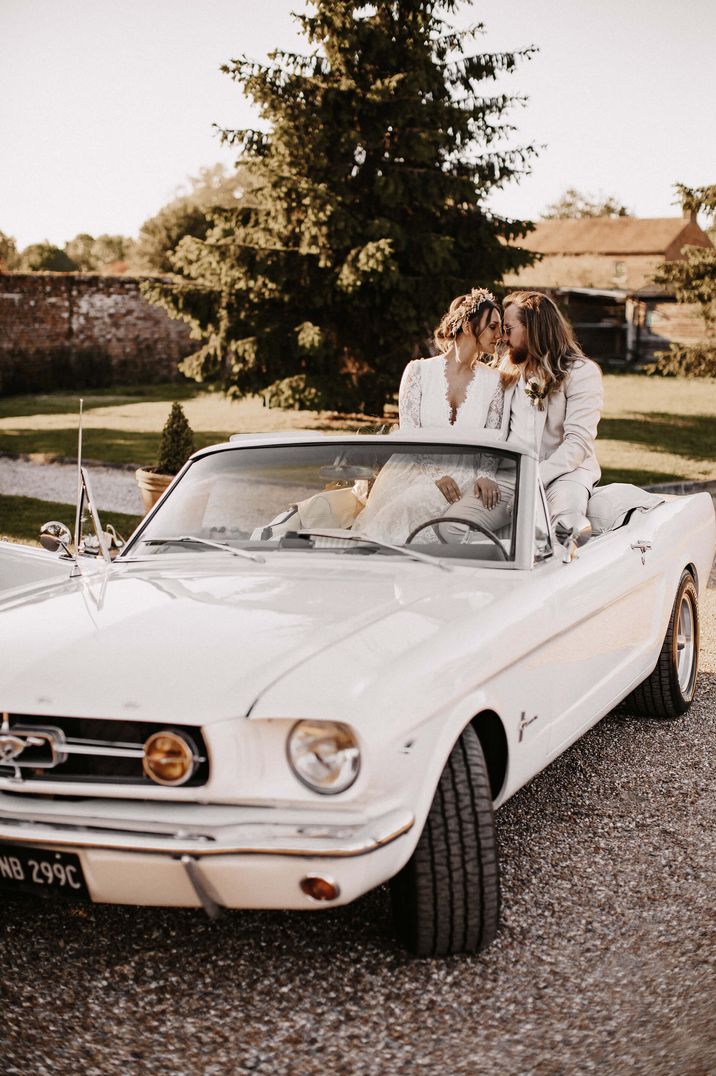 Bride in dried flower crown and groom in white suit in classic white convertible wedding car