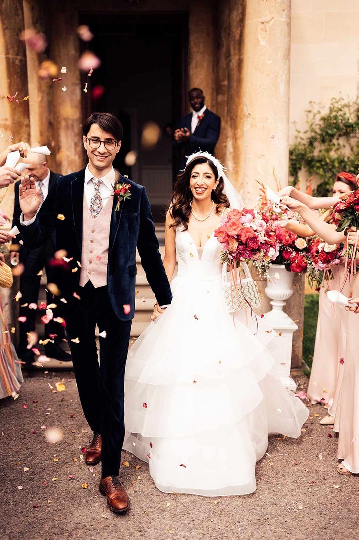 Bride in feathered headband and layered tulle wedding dress holding beaded bag and pink, white and red bridal bouquet walks through petal confetti with groom in dark suit with pink waistcoat