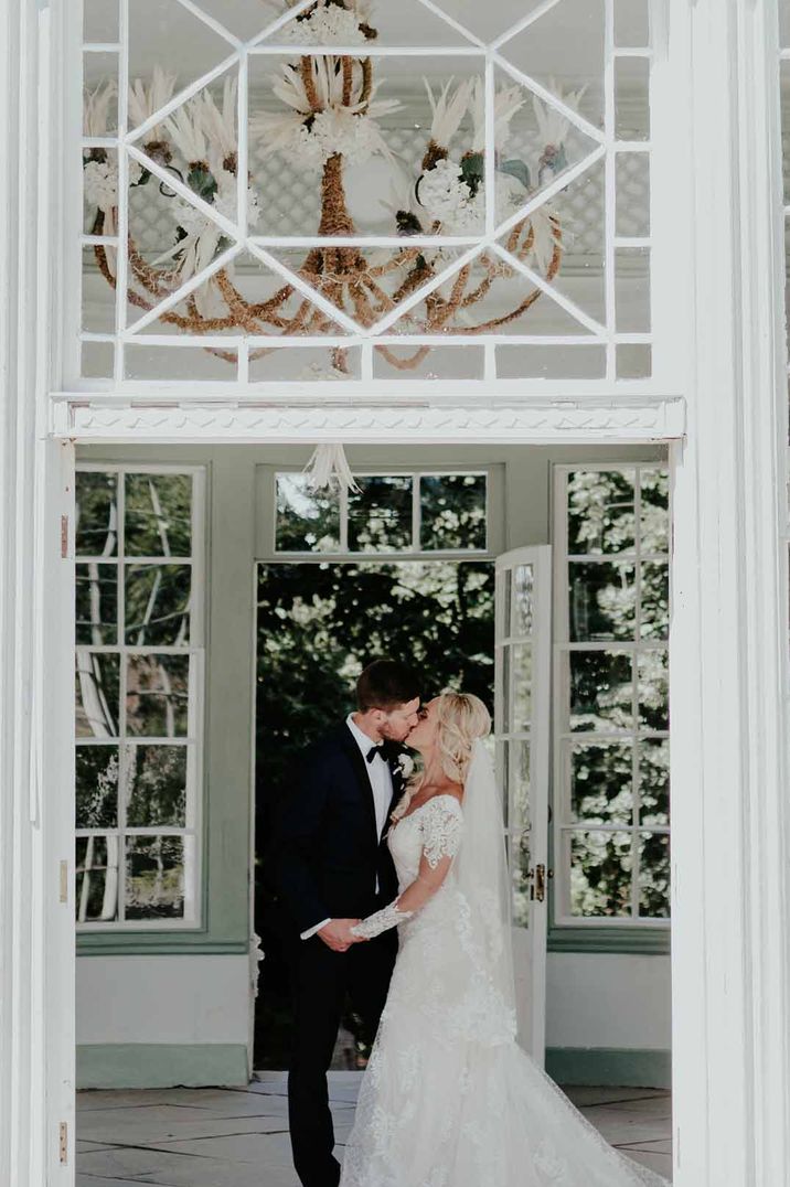 Bride and groom kissing underneath golden chandelier in reception room of Port Eliot Estate glasshouse wedding venue, Cornwall 