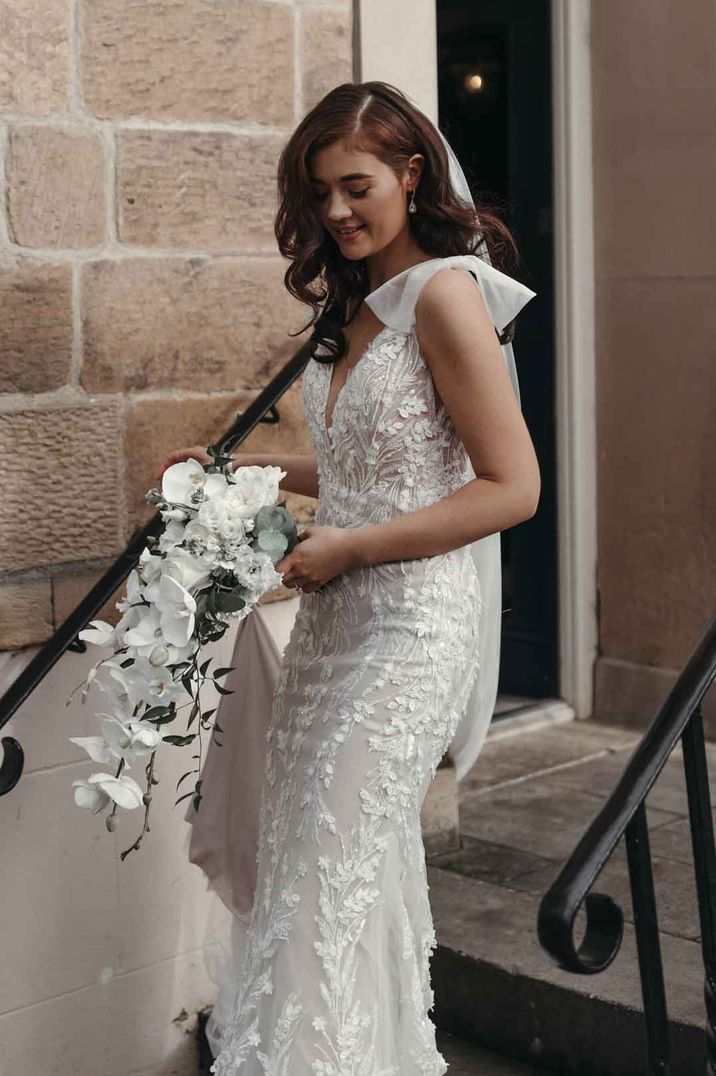 Bride in v neck embellished wedding dress with sheer bow sleeve and white orchid and foliage bouquet 