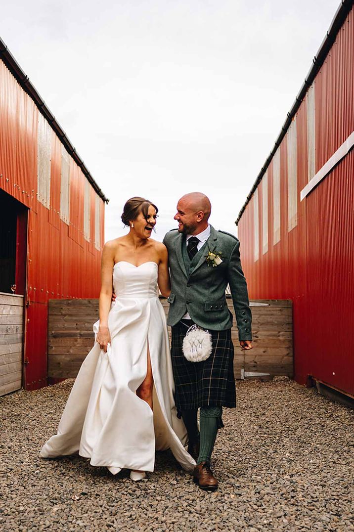 Bride in strapless satin wedding dress with front slit walking with groom in wedding kilt and sporran at Dalduff Luxury Barn Weddings Scotland