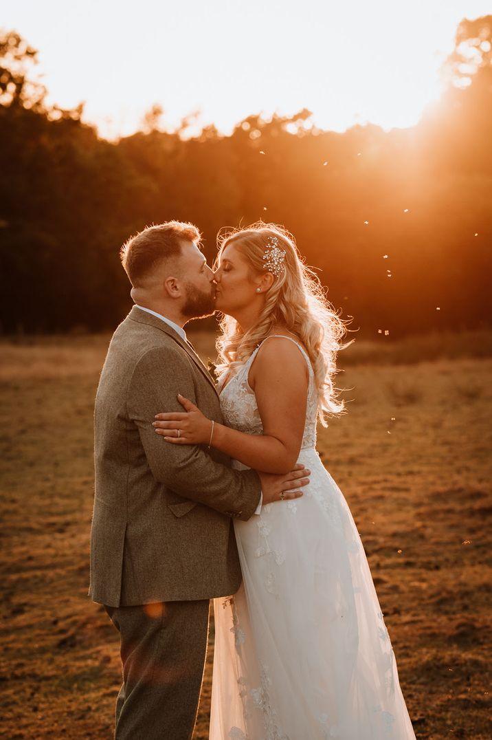 Bride in tulle princess style wedding dress kissing groom in grey suit during warm golden hour 