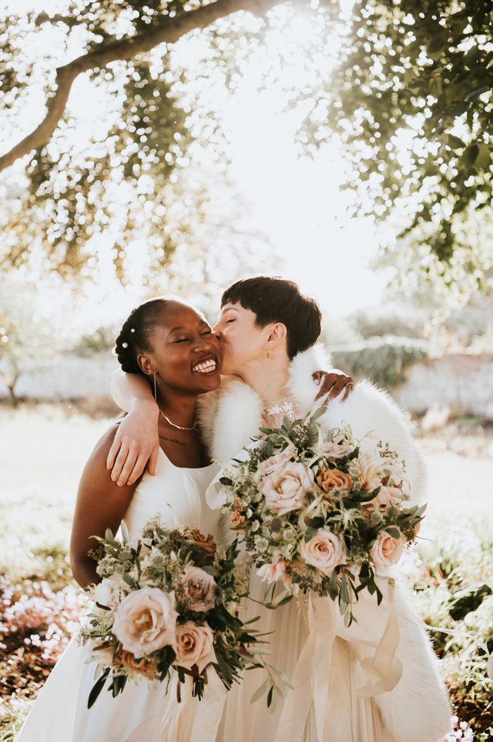 Bride with short hair kissing her bride under a tree at Elmore Court holding romantic pink wedding bouquets 