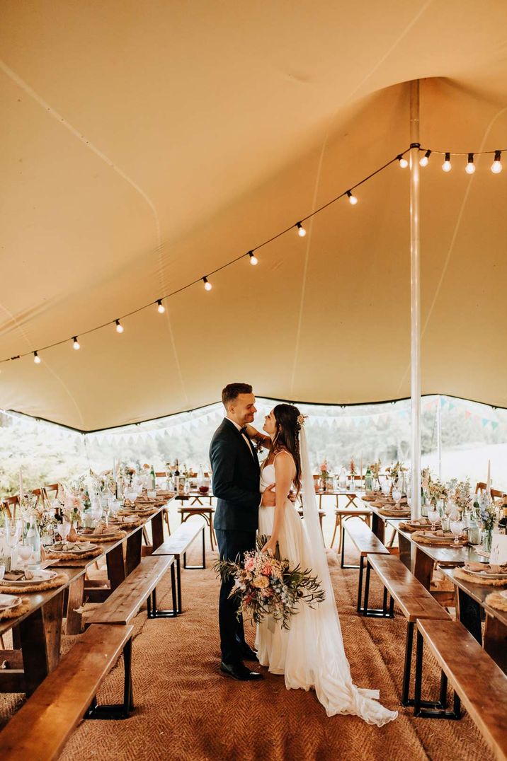 Bride in sleeveless wedding dress with long veil and large neutral bouquet and groom in dark suit standing in DIY tent at garden wedding